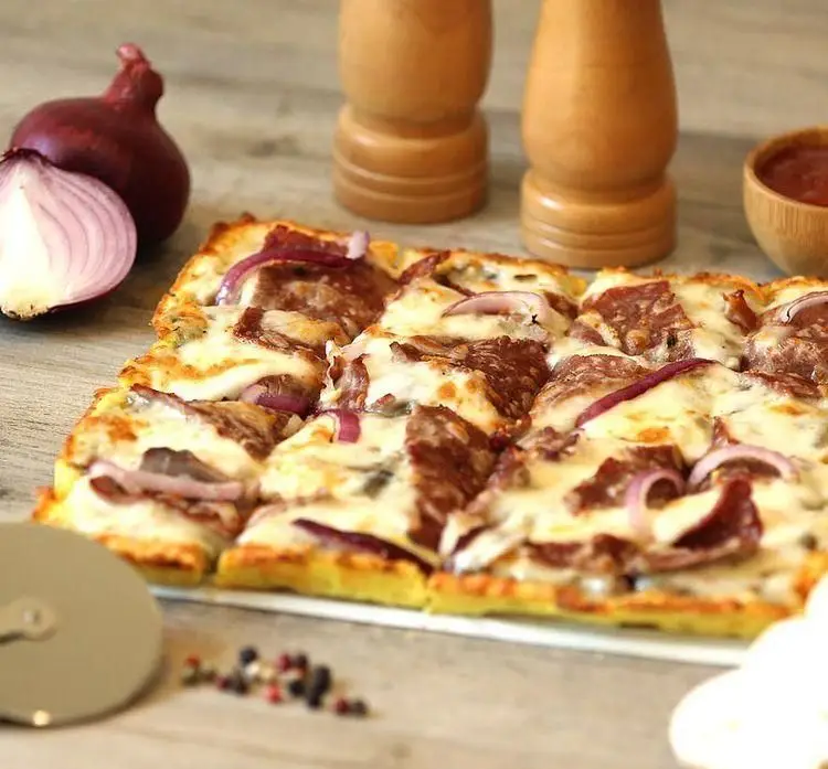 پیتزا پرپروک مستطیلی با برش‌های مربعی، روی میز چوبی، شامل پنیر، پیاز قرمز و کالباس، همراه با پیاز و فلفل‌پاش.