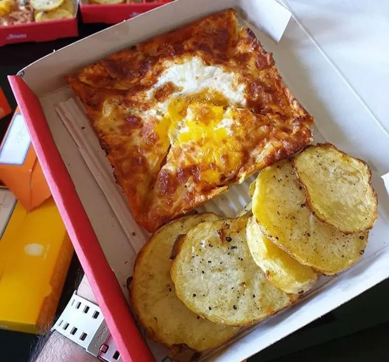 یک وعده غذایی خوشمزه از پیتزا پرپروک، شامل پیتزایی مربعی با تخم‌مرغ نیمرو و سیب‌زمینی‌های برشته و ادویه‌دار در جعبه.