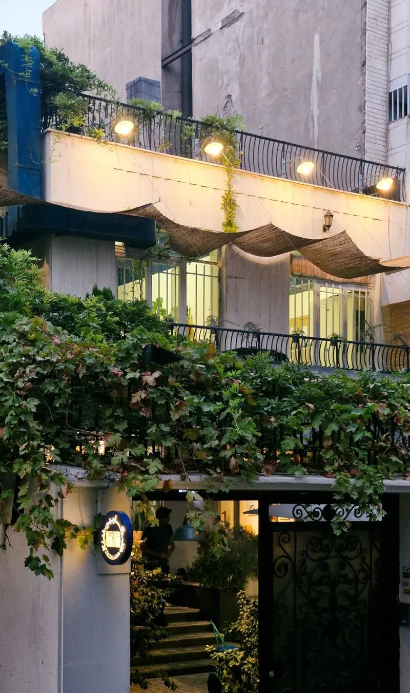 کافه و فضای کار کاربن: نمایی جذاب از ورودی سرسبز، تراس دنج با نورپردازی گرم و فضای کاری الهام‌بخش برای خلاقیت و آرامش.