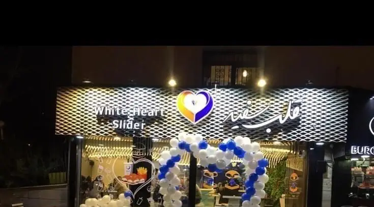 نمای بیرونی رستوران فست فود برگر قلب سفید در شب با تابلوی White Heart Slider و دکوراسیون بادکنکی آبی و سفید برای افتتاحیه.
