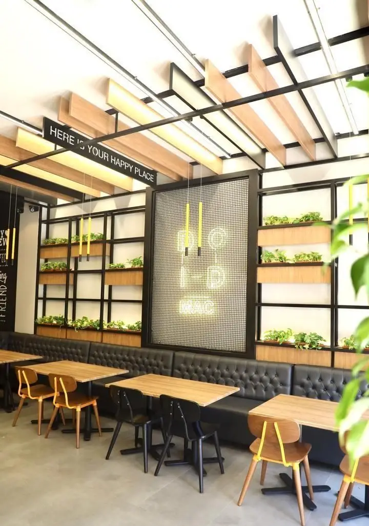 فضای داخلی دنج و مدرن کافه و فست فود دیدومک با سقف چوبی، گلدان‌ها، میز و صندلی‌های شیک و مبلمان راحتی مشکی. اینجا مکان شاد شماست!