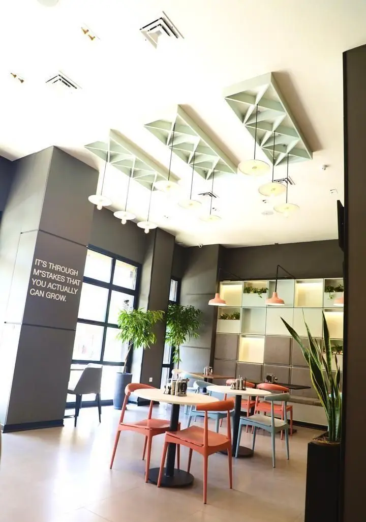 طراحی داخلی چشم‌نواز کافه و فست فود دیدومک. سقف با پنل‌های هندسی و نورپردازی آویز، میز و صندلی رنگارنگ و دیوار سبز دکوری.