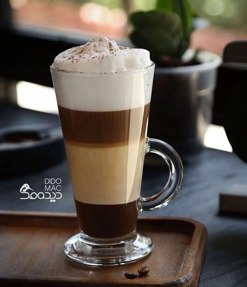 یک لیوان بلند قهوه لایه‌ای زیبای کافه دیدومک، شامل لاته ماکیاتو با فوم و پودر کاکائو، روی سینی چوبی. نوشیدنی خاص و خوشمزه.