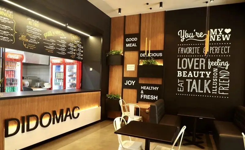 طراحی داخلی شیک و مدرن کافه فست فود دیدومک با پیشخوان چوبی روشن، منو روی تخته سیاه، دیوار تزئینی با جملات مثبت و فضای غذاخوری دلنشین.