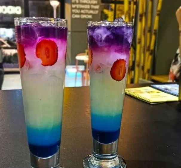 دو نوشیدنی لایه ای رنگی بنفش، صورتی، سبز، آبی با توت فرنگی و یخ در کافه و فست فود دیدومک، انتخابی خنک و جذاب.