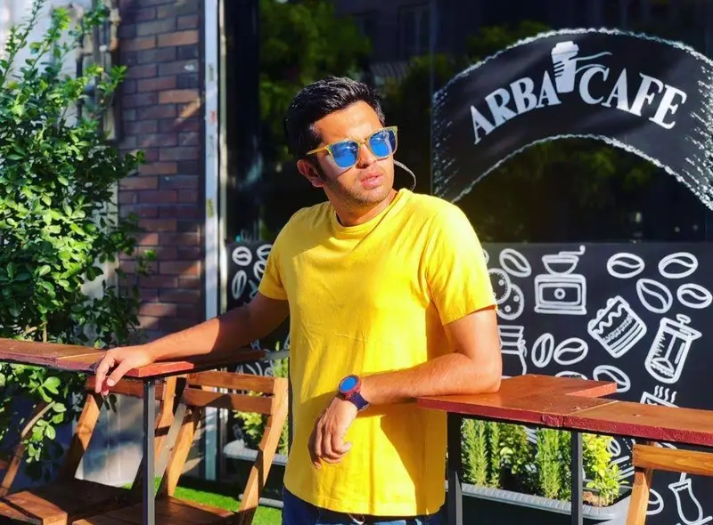 تصویری جذاب از مردی در تی‌شرت زرد و عینک آفتابی، ایستاده در فضای دلنشین کافه آربا با لوگوی مشخص و طرح‌های قهوه روی شیشه.