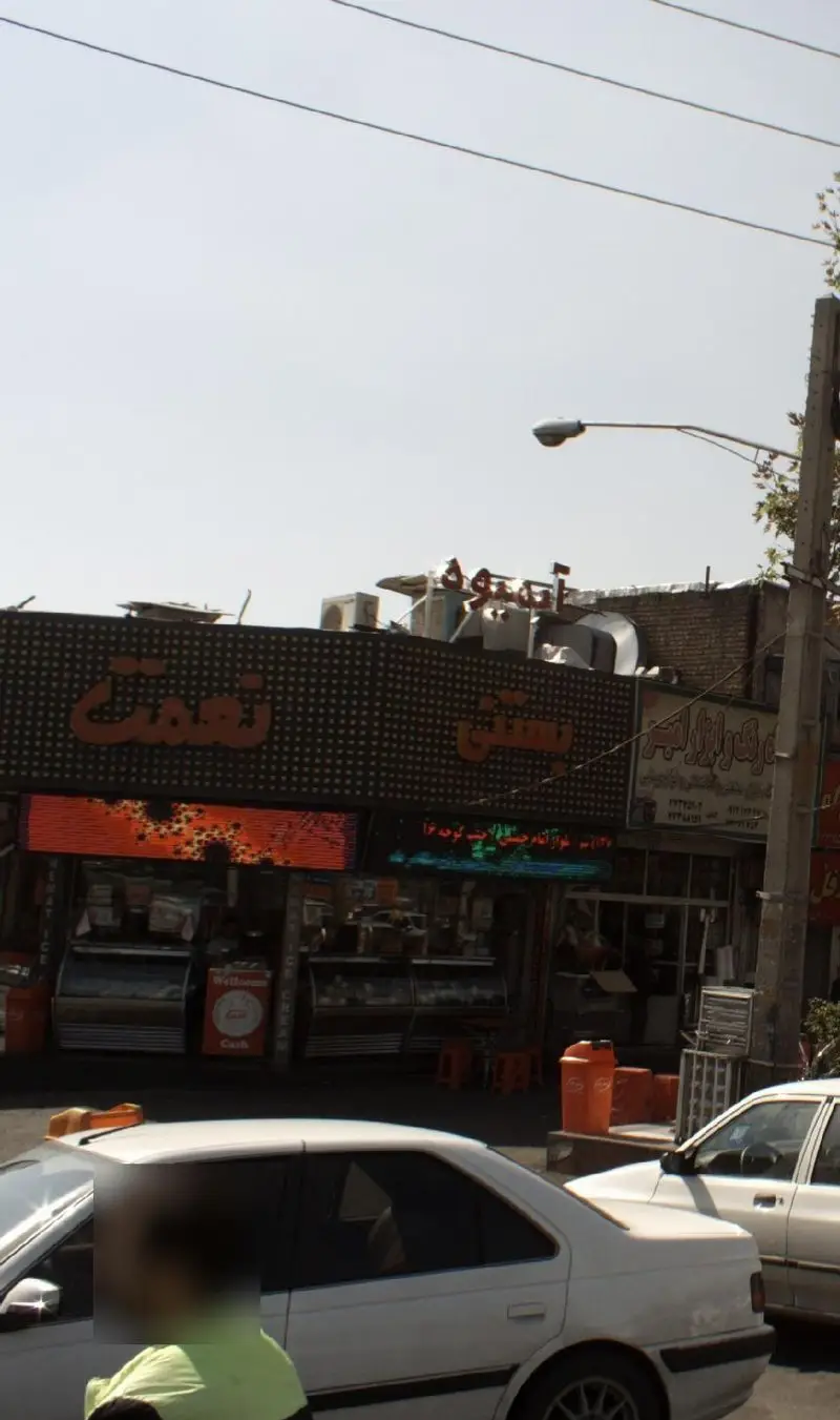 نمای خارجی شعبه بستنی نعمت در یک روز آفتابی، با تابلوهای روشن، ویترین‌های محصولات و خودروها در کنار خیابان شلوغ.