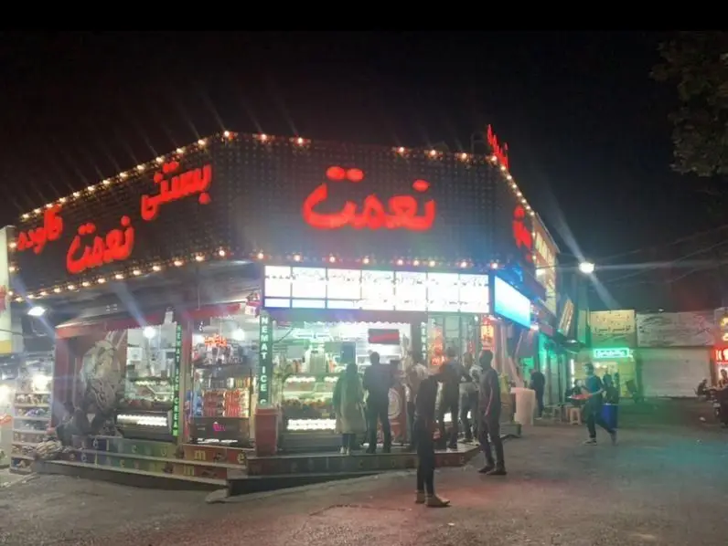 تصویر شب از شعبه بستنی و فالوده نعمت، با تابلوهای نئونی درخشان و مشتریانی در حال خرید در یک خیابان شلوغ