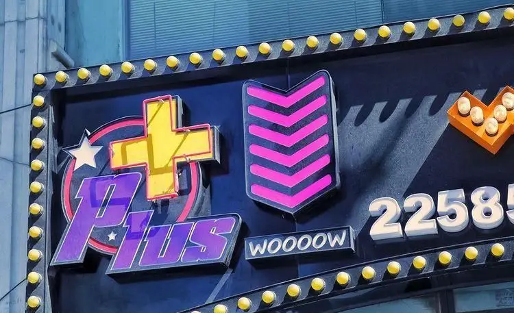 تابلوی فست فود پلاس با لوگوی پلاس زرد، حروف بنفش، ستاره، فلش‌های صورتی و حاشیه لامپ برای جلب توجه مشتریان در شب.