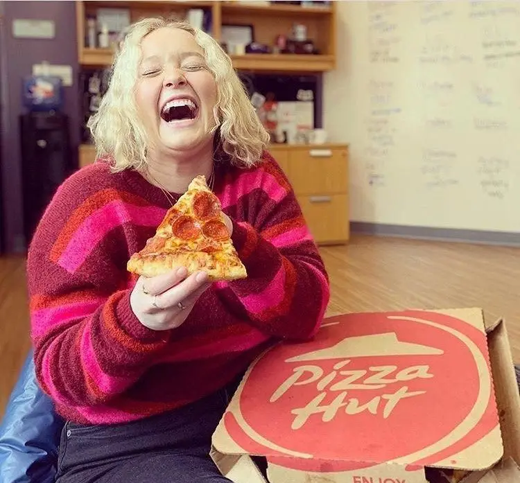 زن جوان با موهای بلوند در حال خنده و لذت بردن از پیتزا هات اکباتان. او یک برش پیتزا پپرونی تازه و خوشمزه در دست دارد.