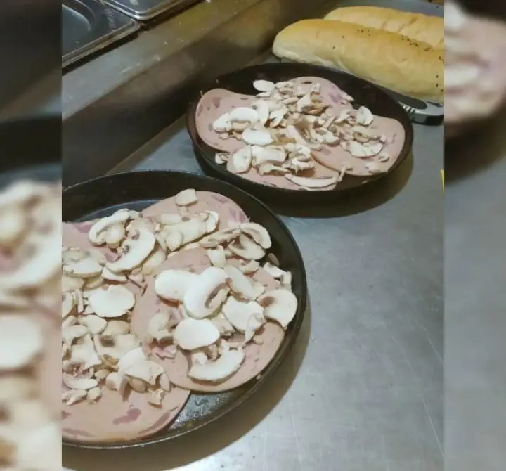 صحنه‌ای از آماده‌سازی غذا در پیتزا هات اکباتان: دو ظرف حاوی کالباس ورقه‌ای و قارچ اسلایس شده، با نان‌های باگت تازه در پس‌زمینه.