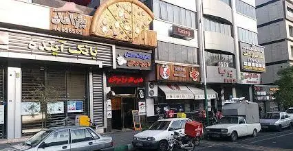 تصویری از نمای بیرونی پیتزا هات اکباتان در یک خیابان پرتردد با چندین کسب و کار و تابلوهای تبلیغاتی.