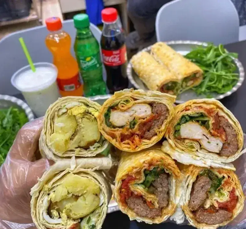 تصویری جذاب از لقمه‌های لذیذ رستوران لقمه بازار: ساندویچ‌های رول کباب، مرغ و سیب‌زمینی، در کنار انواع نوشیدنی و سبزی تازه. تجربه‌ای خوشمزه.