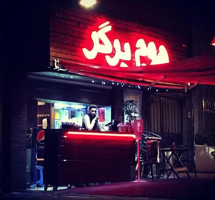 نمای شبانه رستوران هوم برگر با تابلوی نئون قرمز روشن، پیشخوان پذیرایی و کارکنان داخل، همراه با فضای دلنشین و میزهای بیرونی.