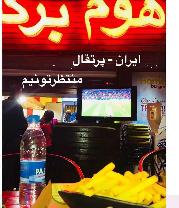 مشتریان در رستوران هوم برگر مشغول تماشای فوتبال ایران و پرتغال، همراه با سیب زمینی سرخ کرده و نوشیدنی. فضایی هیجان‌انگیز برای لذت بردن از مسابقه.