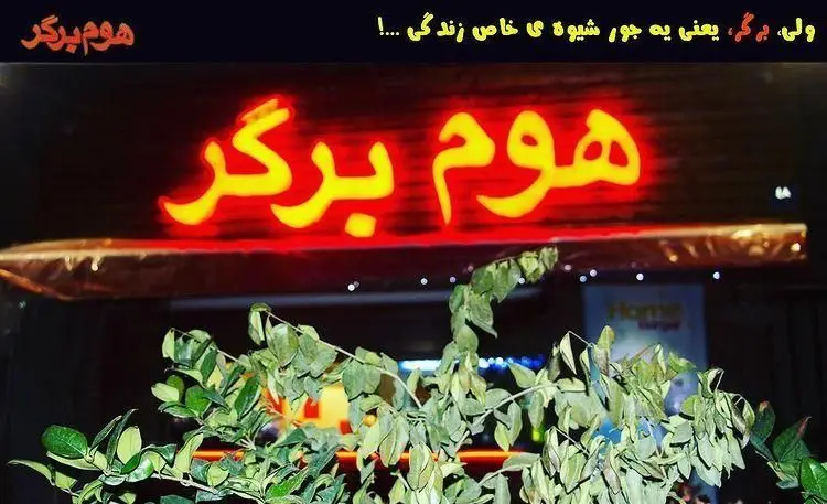 تابلو نئون زرد و قرمز فست فود "هوم برگر" (Home Burger) در شب، با شعار "یعنی یه جور شیوه ی خاص زندگی...!" در بالای آن. تصویری از نمای بیرونی.