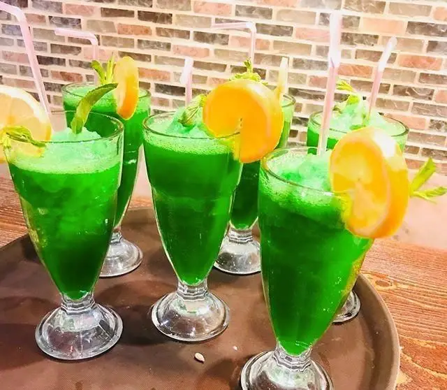 چندین لیوان نوشیدنی اسلاشی سبز خنک با تزئین پرتقال و نعنا، سرو شده در کافی شاپ ترمه، انتخابی گوارا برای تابستان.