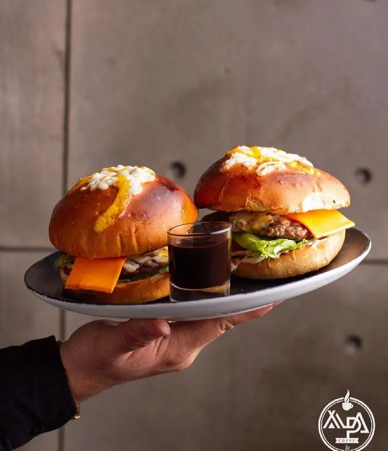 دو برگر ویژه و لذیذ با نان پرزل و پنیر آب شده، همراه با سس مخصوص، در بشقاب مشکی کافه رستوران آلپا سرو شده است.