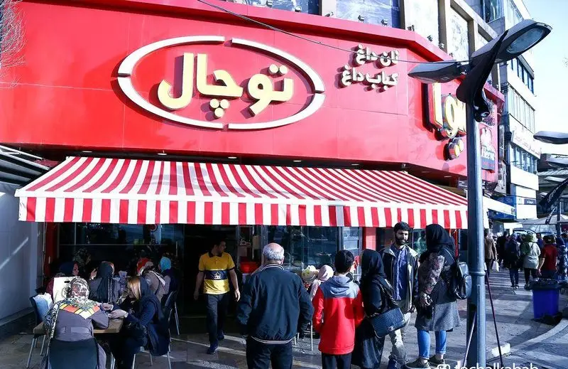 نمای بیرونی کباب سرای توچال با تابلو قرمز بزرگ، سایبان راه راه قرمز و سفید و مشتریان در حال صرف کباب داغ و نان داغ.