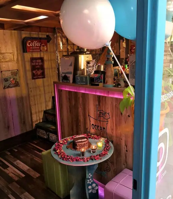 فضای داخلی کافه اوشن با دکوراسیون چوبی، میز تولد شامل کیک، شمع و گلبرگ، بالون و تابلوهای قهوه. مکانی دنج برای جشن و قرارهای عاشقانه.