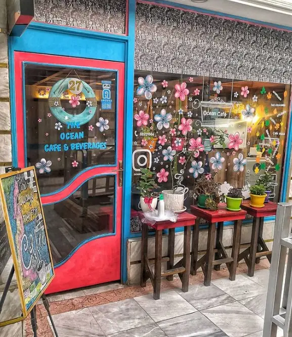 نمای بیرونی کافه و نوشیدنی‌های اوشن، با طراحی رنگارنگ درب آبی قرمز و پنجره‌های گلدار، و تابلوی قهوه، مکانی دنج و زیبا.