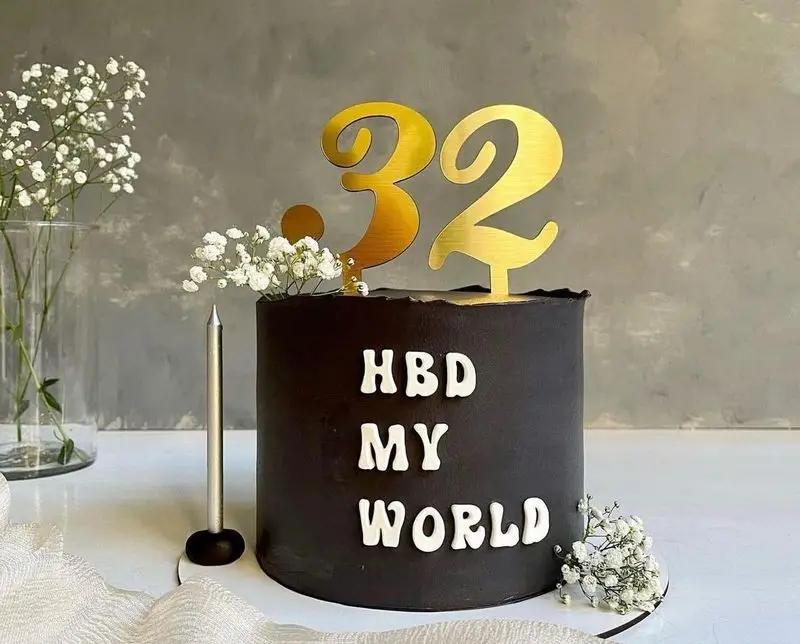 کیک تولد مشکی لاکچری میس کیک، با تاپر طلایی عدد ۳۲ و متن HBD MY WORLD. سفارش آنلاین کیک با دیزاین گل عروس.