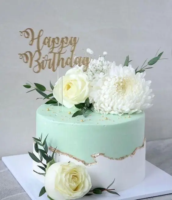 سفارش کیک تولد لوکس از میس کیک: کیک دو طبقه سبز آبی و سفید با گل آرایی طبیعی، تاپر Happy Birthday و حاشیه طلایی زیبا.