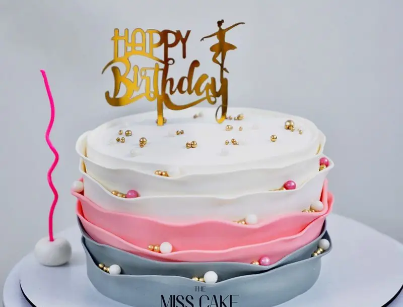 کیک تولد دخترانه زیبا و خاص از میس کیک. طراحی چند طبقه رفل با رنگ‌های سفید، صورتی و طوسی. تاپر بالرین هپی برثدی.