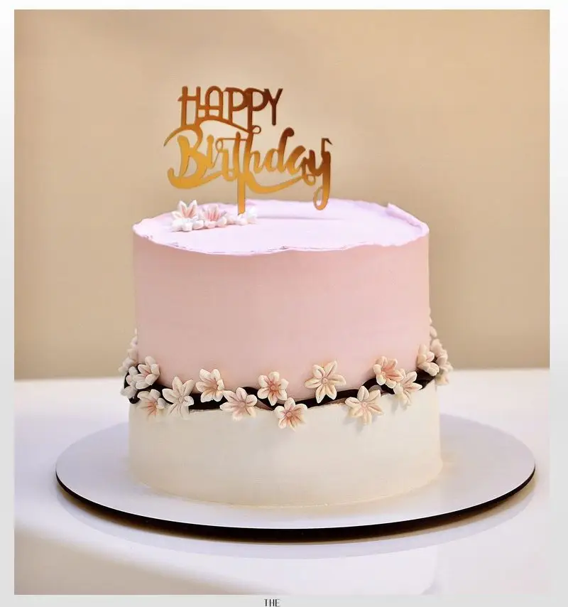 سفارش کیک تولد صورتی و سفید میس کیک با تاپر طلایی Happy Birthday و تزیین هنرمندانه گل‌های ظریف فانتزی مناسب جشن و مراسم.