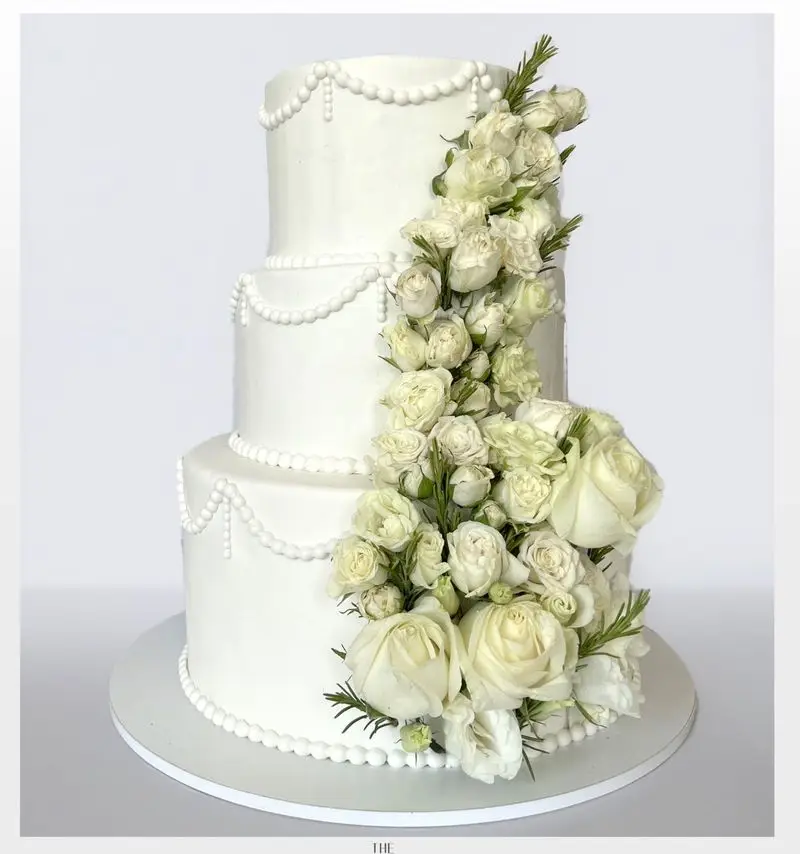 کیک عروسی سه طبقه سفید خیره‌کننده میس کیک با دیزاین خاص رزهای سفید آبشاری و مروارید. مناسب برای سفارش کیک و شیرینی لوکس.
