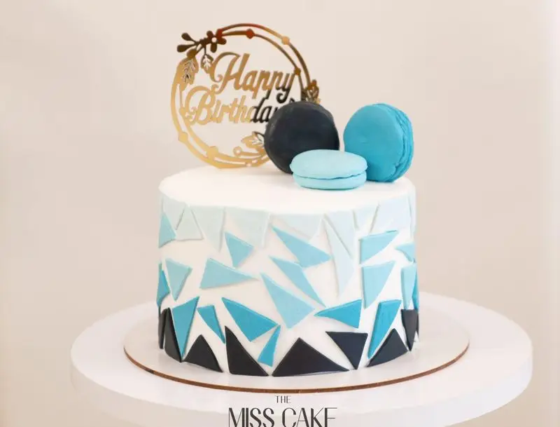 سفارش کیک تولد مردانه/پسرانه آبی با طرح هندسی مثلثی گرادیان، مزین به ماکارون و تاپر هپی برس دی از میس کیک.