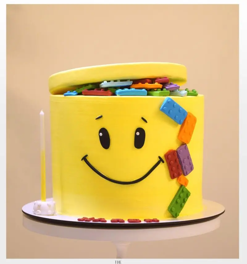 کیک تولد زرد و شاد میس کیک با صورتک خندان و تزئین لگو رنگی، ایده‌آل برای سفارش کیک بچگانه و جشن تولد با شمع.