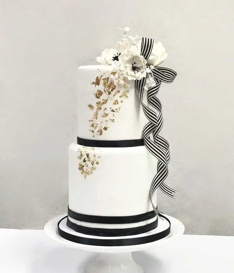 کیک دو طبقه سفید و شیک میس کیک با تزیین برگ طلایی، گل شکری سفید و روبان راه راه مشکی سفید، مناسب جشن و سفارش.