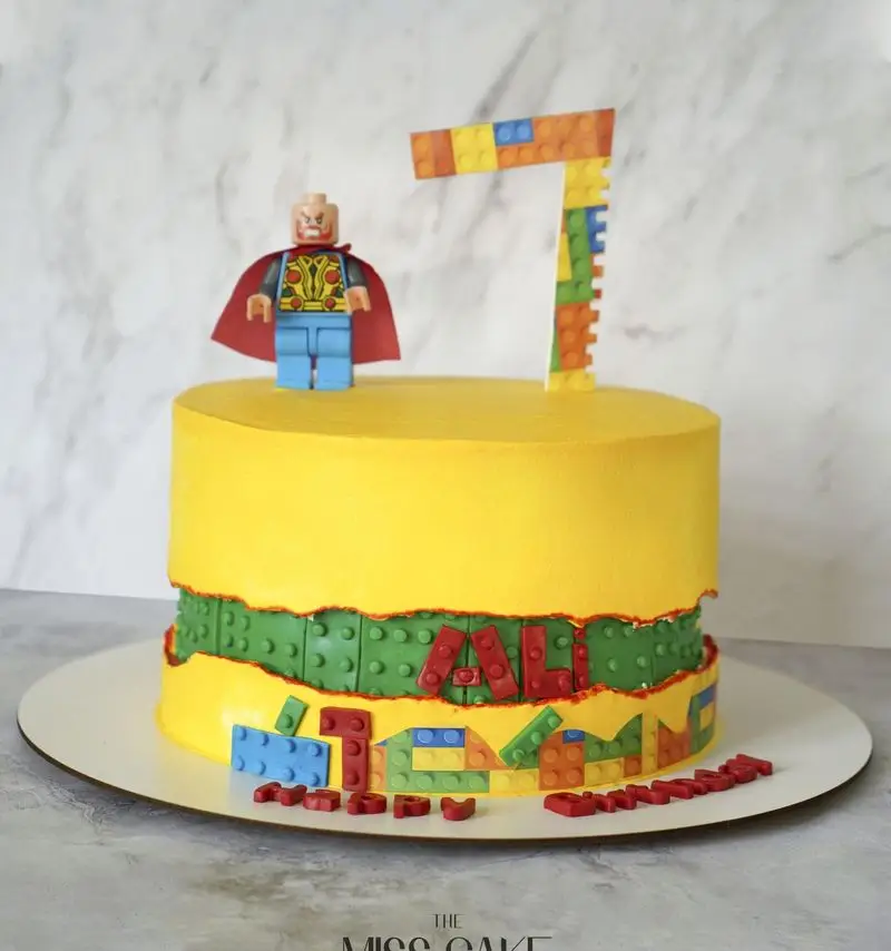 کیک تولد لگو پسرانه زرد رنگ با تم ابرقهرمانی، فیگور لگو ثور، عدد ۷ لگویی و نام "علی". سفارش کیک خاص میس کیک.