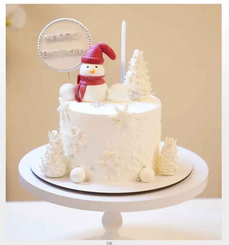 کیک تولد با تم زمستانی میس کیک: کیک سفید با تزیین آدم برفی، کلاه و شال قرمز، درخت کریسمس، دانه برف و شمع.