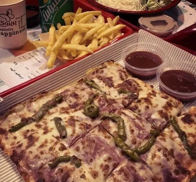 نمایی نزدیک از یک وعده غذایی کامل در پیتزا پرپروک: پیتزای مربع با فلفل و پنیر، سیب‌زمینی سرخ‌کرده، نوشابه و سس‌های مخصوص.