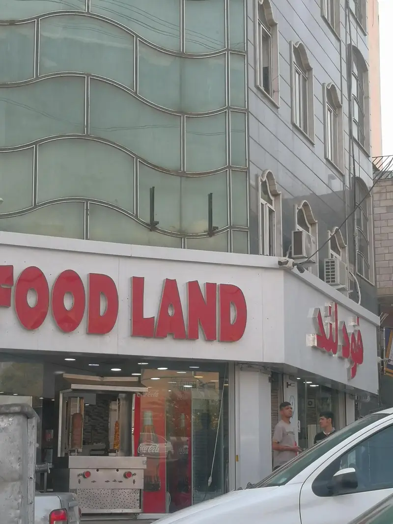تصویر نمای بیرونی رستوران و فست فود فودلند با تابلوی FOOD LAND و فودلند به دو زبان، دستگاه شاورما در ورودی و معماری مدرن ساختمان.