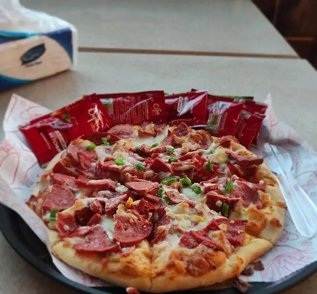 پیتزای خوشمزه با تکه‌های کالباس، پنیر ذوب‌شده و فلفل سبز، سرو شده در کافه و رستوران عمارت، همراه با بسته‌های سس کچاپ.