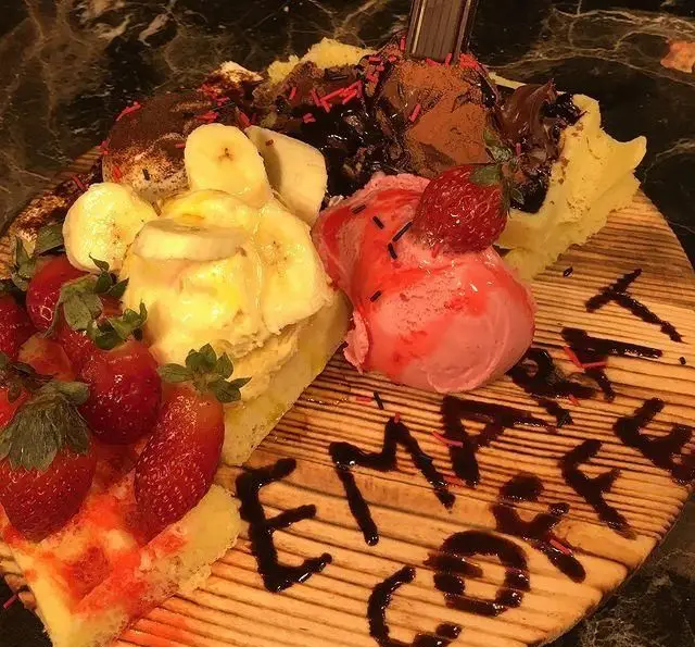 بشقاب دسر جذاب کافه و رستوران عمارت شامل وافل، توت فرنگی، موز، بستنی توت فرنگی و شکلاتی با سس شکلات روی تخته چوبی.