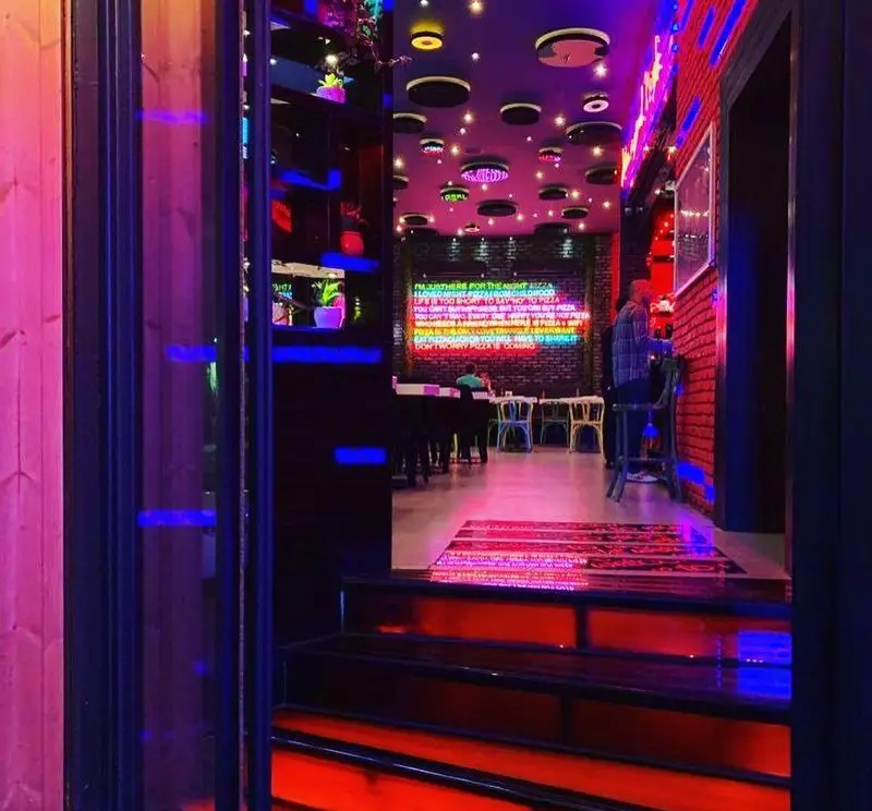 فضای داخلی مدرن و رنگارنگ پیتزا شب با نورپردازی نئونی قرمز و آبی، سقف خلاقانه، دیوار آجری و محیطی ایده‌آل برای پیتزا‌ دوستان.