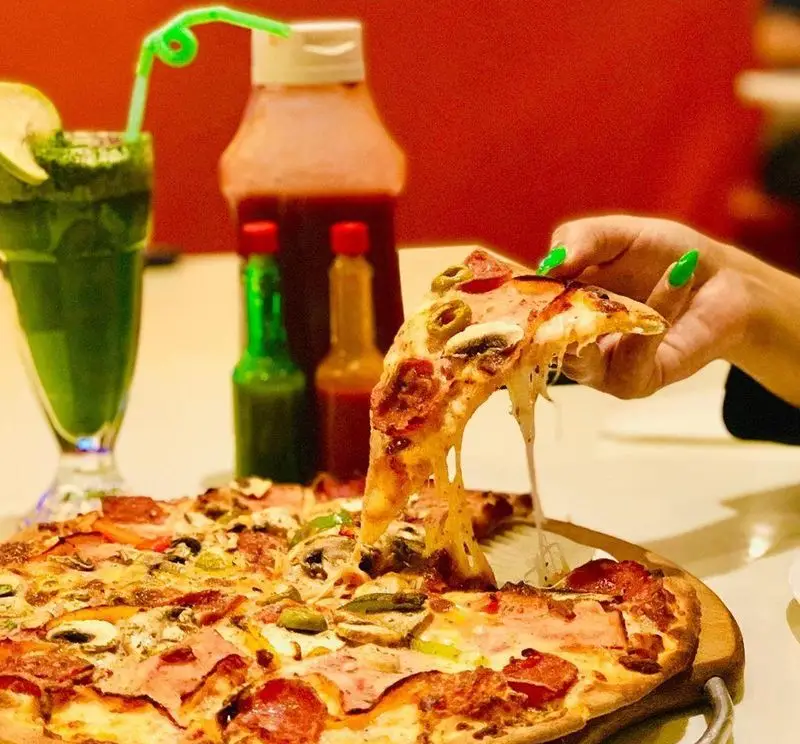 تصویری جذاب از پیتزا شب: یک برش پیتزا لذیذ با پنیر فراوان کش‌دار، پپرونی، قارچ و زیتون، کنار نوشیدنی سبز خنک و سس‌ها.