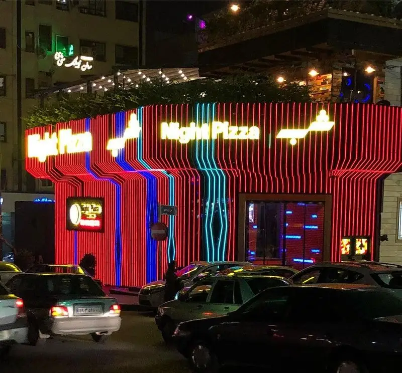 نمای مدرن فست فود پیتزا شب با نورپردازی نئون خیره‌کننده قرمز و آبی در یک شب پر رفت و آمد.