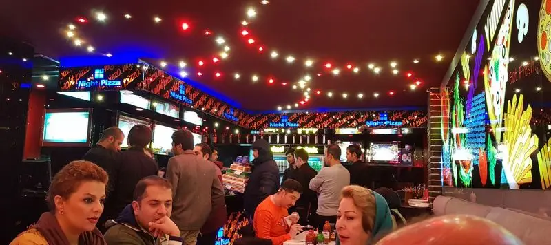 فضای داخلی پرجنب‌وجوش و دلپذیر پیتزا شب. مشتریان در حال لذت بردن از پیتزا در محیطی با دکوراسیون مدرن، نورپردازی رنگارنگ و طرح‌های غذایی جذاب.