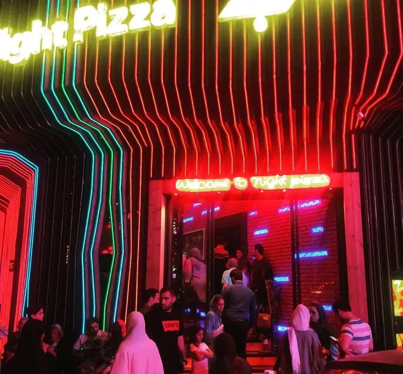 نمای خیره‌کننده پیتزا شب با تابلوی نئون زردرنگ و نورپردازی‌های خطی قرمز، آبی و سبز، به همراه ورودی پر جنب و جوش در شب.