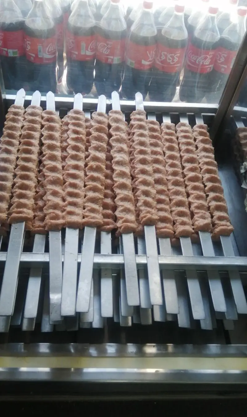 کباب کوبیده تازه و آماده پخت تهیه غذای امیر در سیخ‌های منظم، همراه با بطری‌های کوکاکولا در پس‌زمینه