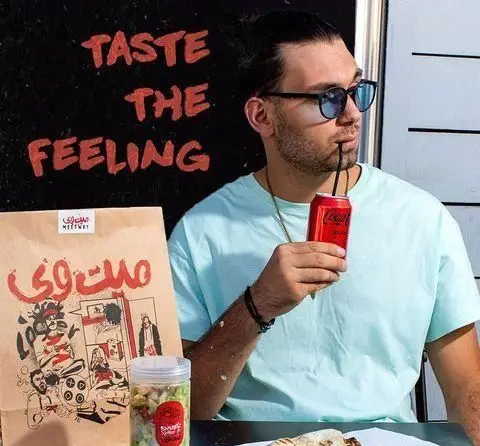 مردی با ساندویچ و نوشیدنی کوکاکولا از غذای میت وی لذت میبرد. بستهبندی جذاب و شعار "TASTE THE FEELING" در پسزمینه.