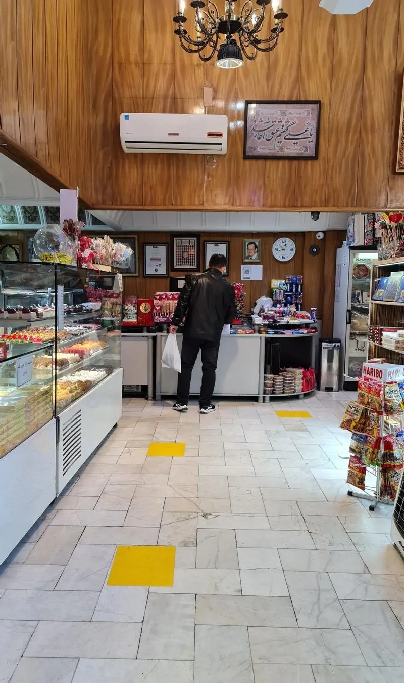 نمای داخلی شیرینی فروشی سوگل، با ویترین‌های پر از شیرینی و دسر تازه. مشتری در حال خرید. محیطی دلنشین برای عاشقان شیرینی و کیک.