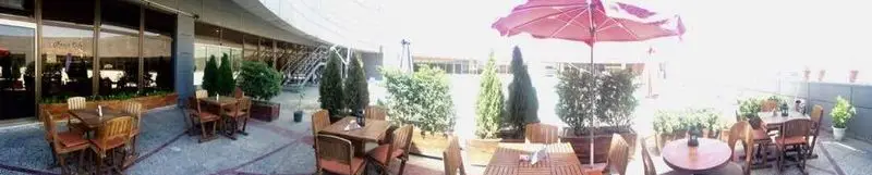 فضای بیرونی دنج و زیبای نسکو کافه با میز و صندلی‌های چوبی، چتر بزرگ قرمز، گلدان‌های سبز و نمایی از ساختمان مدرن در پس‌زمینه.