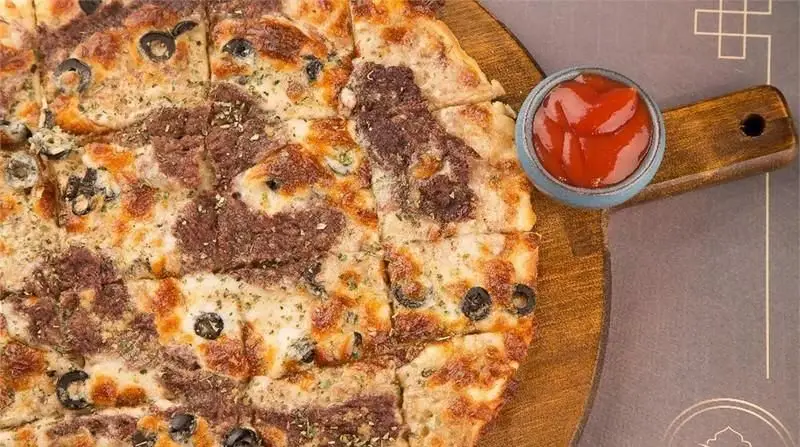 پیتزا مربعی با گوشت چرخ کرده، پنیر ذوب شده و زیتون سیاه، پاشیده شده با سبزیجات معطر، سرو شده روی تخته چوبی در کافه رستوران آشا. سس کچاپ در کنار.