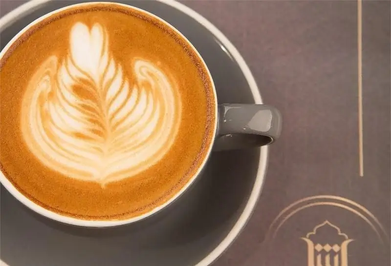 یک فنجان قهوه لاته داغ و خوش‌طعم با لاته آرت زیبای طرح برگ در کافه رستوران آشا، مکانی دلنشین برای سرو نوشیدنی‌های گرم.